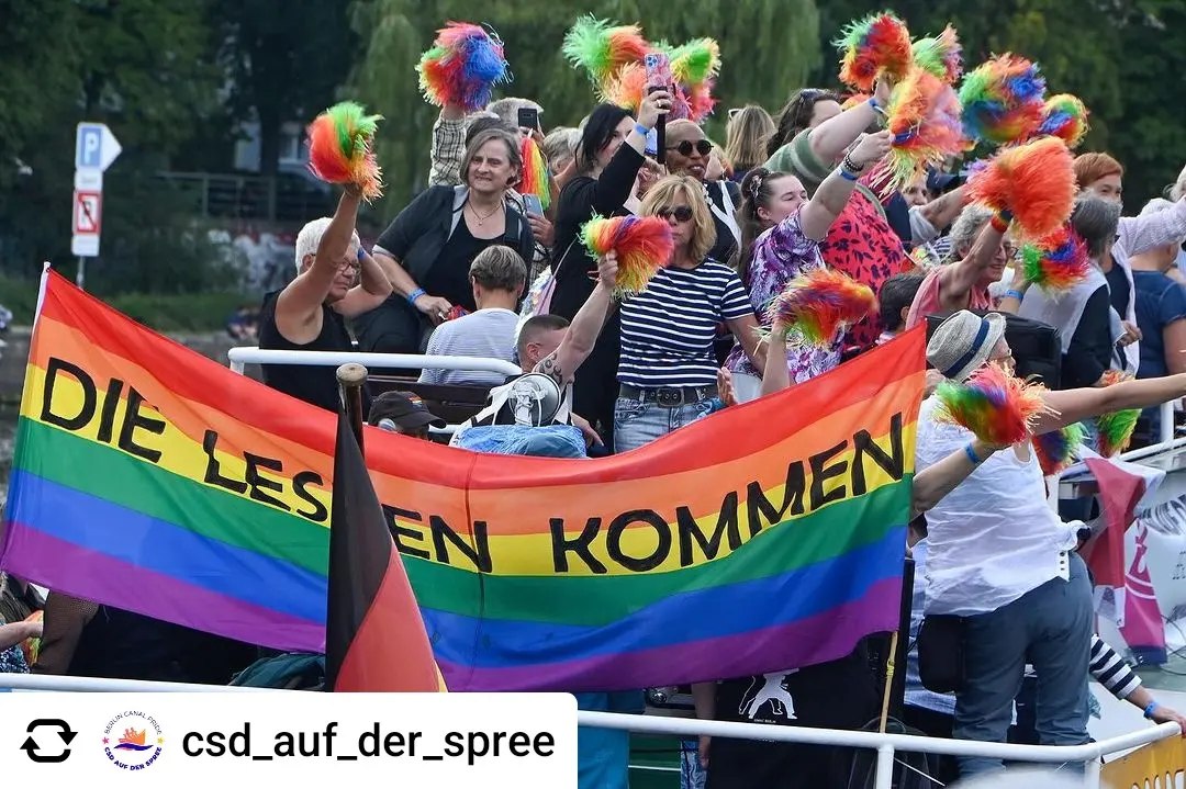 Ein Foto von vielen Frauen auf einem Boot die lachen. Vorne sieht man eine Regenbogenflagge worauf steht: Lesben kommen 