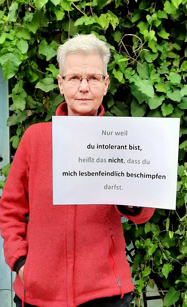 Ein Foto von einer älteren Frau mit kurzem grauen Haar, die einen beschrifteten Zettel vor sich hält.