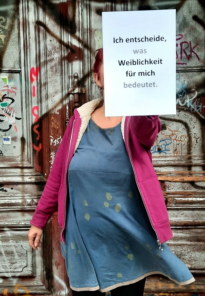 Ein Foto von einer Frau im Kleid, die einen beschrifteten Zettel vor sich hält.