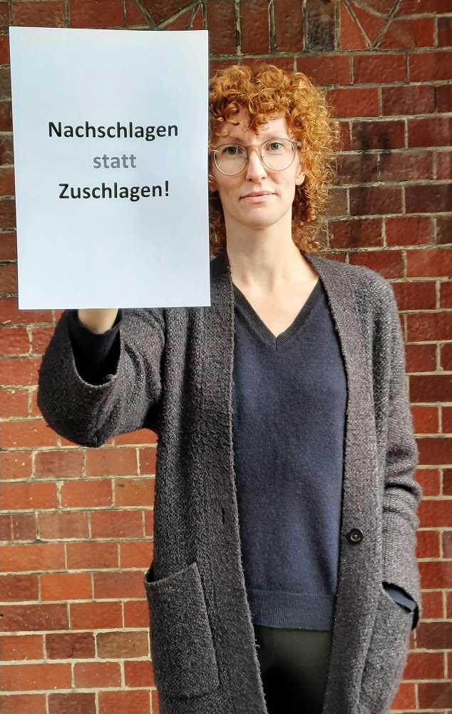 Ein Foto von einer jungen Frau mit Brille, die einen beschrifteten Zettel vor sich hält.