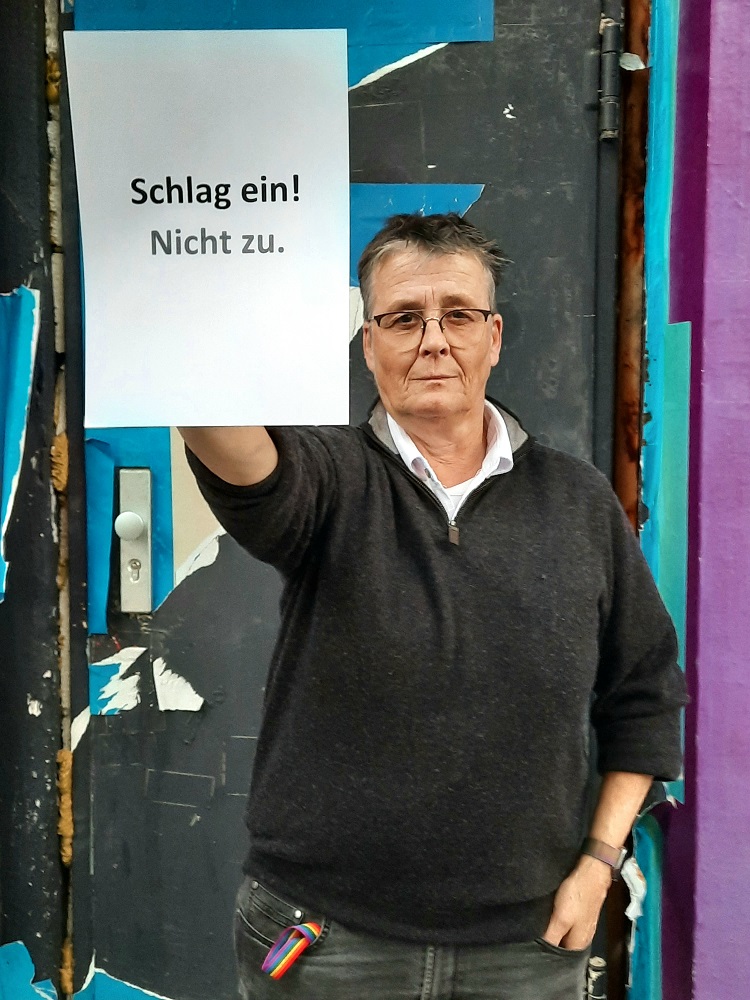 Ein Foto von einer Frau mit kurzem Haar und einer Brille, die einen beschrifteten Zettel vor sich hält.