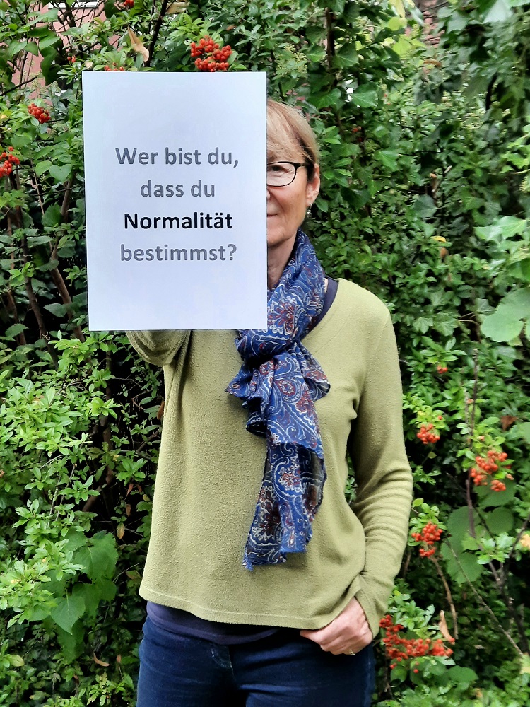 Ein Foto von einer  Frau mit Schaal, die einen beschrifteten Zettel vor sich hält.