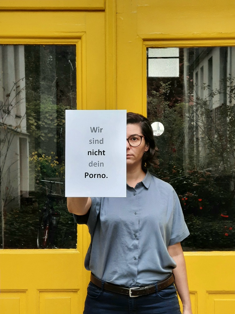 Ein Foto von einer jungen Frau mit Brille, die einen beschrifteten Zettel vor sich hält.